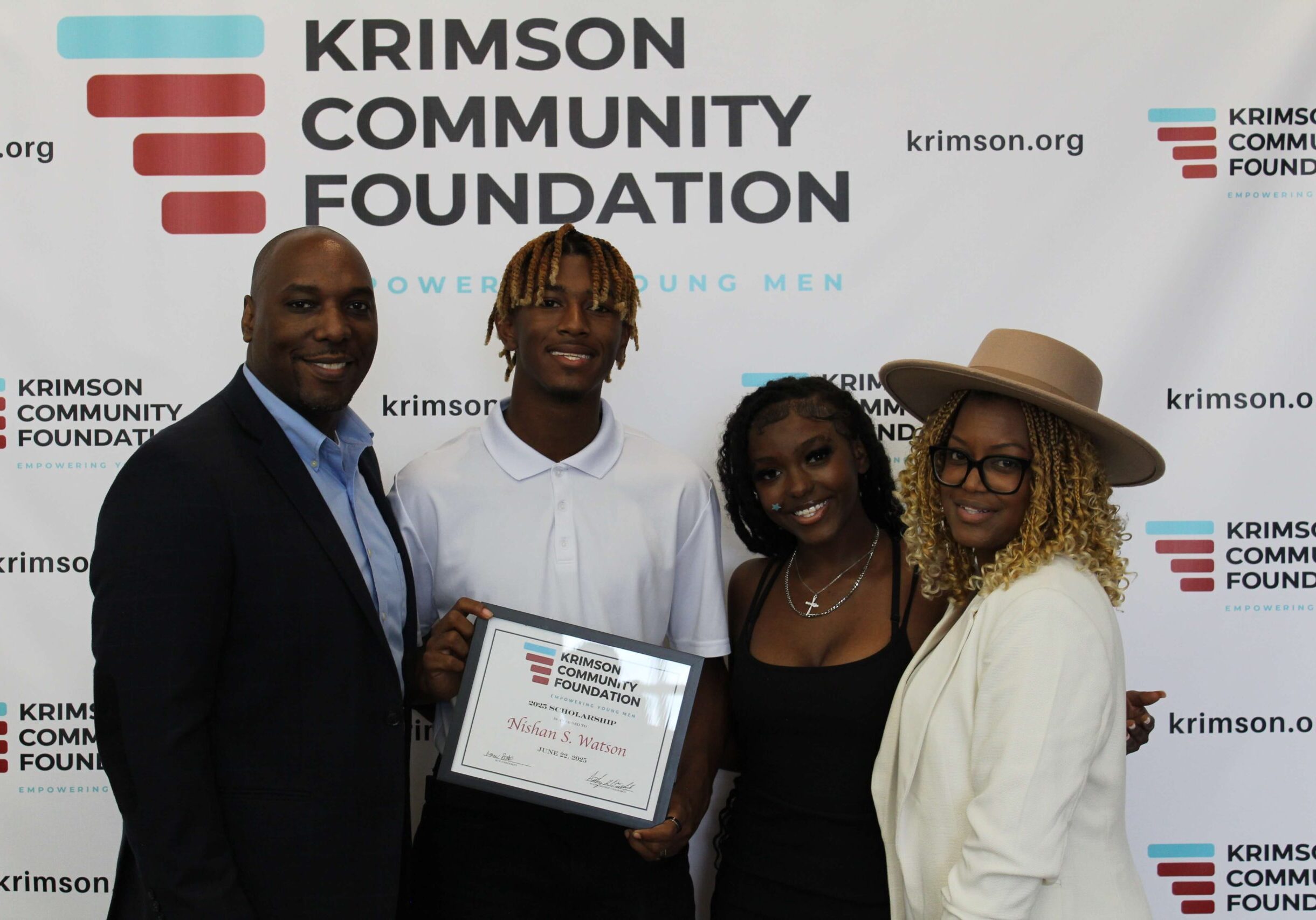 A group of young men holding certificates pose with adults in front of a Krimson Community Foundation scholarship sign in a conference room.